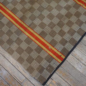 Brown and tan checkered flatweave rug with a diagonal red and gold stripe rests on gray wood planks.