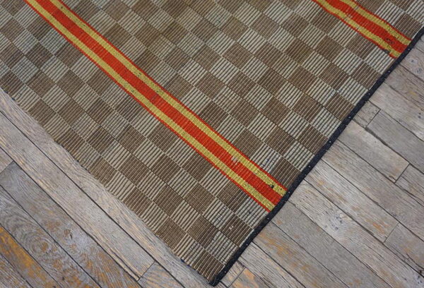 Brown and tan checkered flatweave rug with a diagonal red and gold stripe rests on gray wood planks.