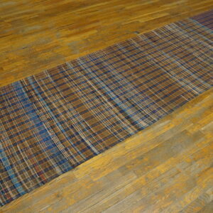A brown and blue plaid American Rag Rug runner with a dark striped end rests on a worn wood floor.