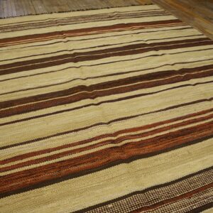 A large flatweave rug with wide beige and thin brown and rust horizontal stripes rests on distressed wood flooring.