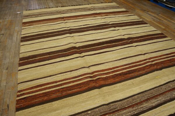 A large flatweave rug with wide beige and thin brown and rust horizontal stripes rests on distressed wood flooring.