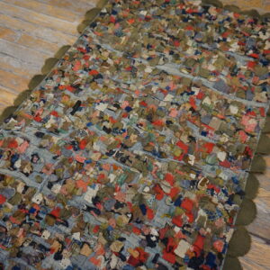 A highly textured, multicolored scrap rug with an olive scalloped edge rests on distressed wood flooring.