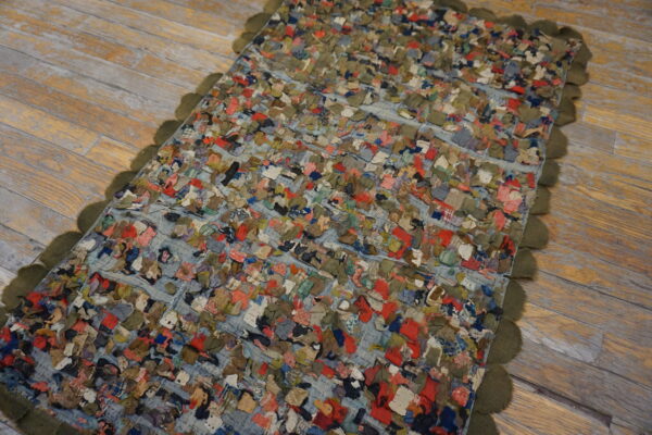 A highly textured, multicolored scrap rug with an olive scalloped edge rests on distressed wood flooring.
