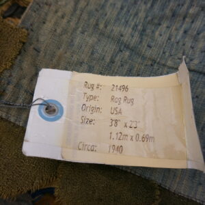 Detail of a coarsely woven dark green and speckled blue textile rug showing a product identification tag attached by wire.