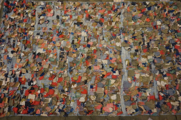 Multicolored, high-texture textile with red, green, and blue scraps stitched over a blue grid pattern.