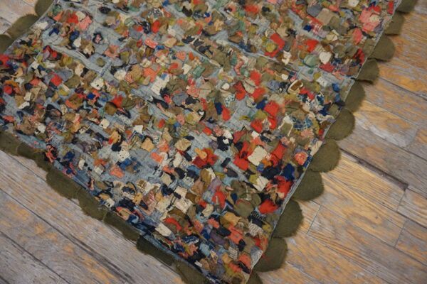 Multicolored fabric scraps create a textured rag rug with an olive scalloped edge on light wood floorboards.
