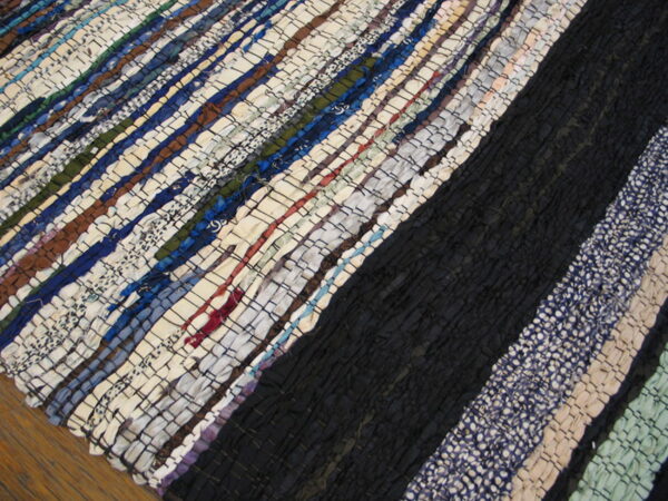 Textured rag rug with wide diagonal stripes in dark blue, black, cream, and multicolored scrap fabrics on wood flooring.