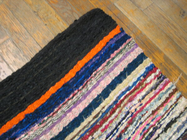 Close-up of a woven striped rug with colorful rag texture and a solid black corner on wood.