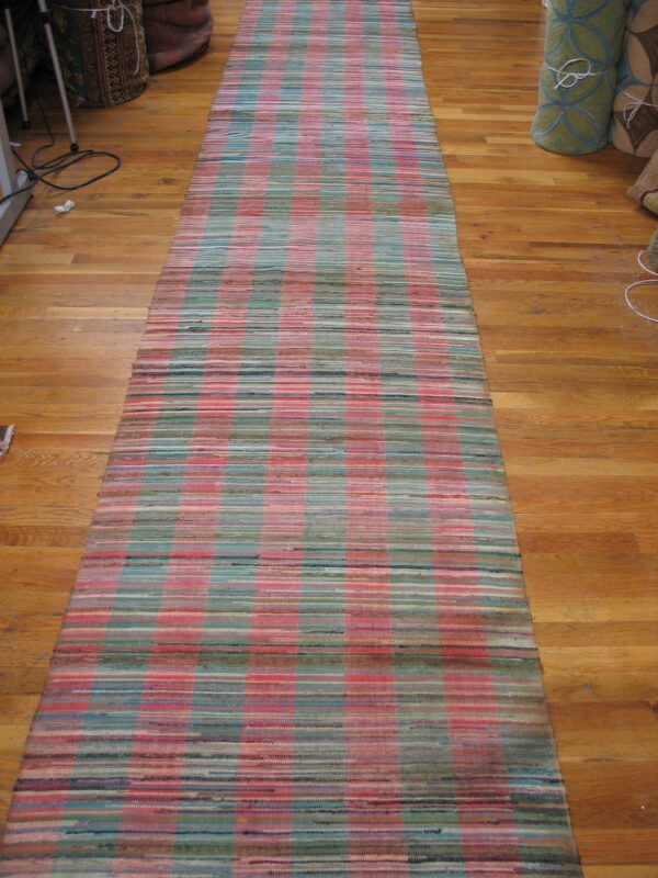 A woven rag rug runner featuring a plaid pattern of pink, teal, and green lies on a wood floor.