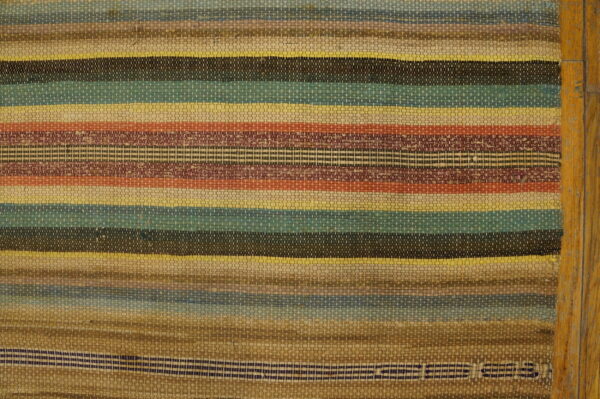 A flat-weave rag rug features horizontal stripes in multicolored shades of brown, teal, yellow, and rust next to wood planks.