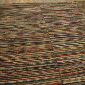 Low pile rug with variegated horizontal stripes in mixed earth tones rests on peeling yellow wood planks.