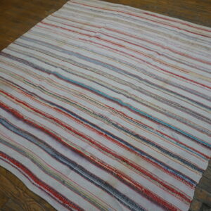 A white flatwoven rag rug with thin colorful stripes rests on dark rustic wood flooring.