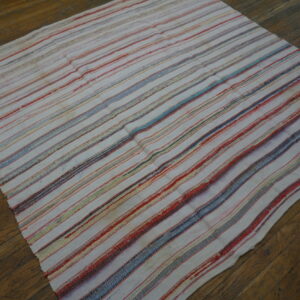 A flat-textured rug with cream, red, and blue stripes lies diagonally on an aged wood floor.
