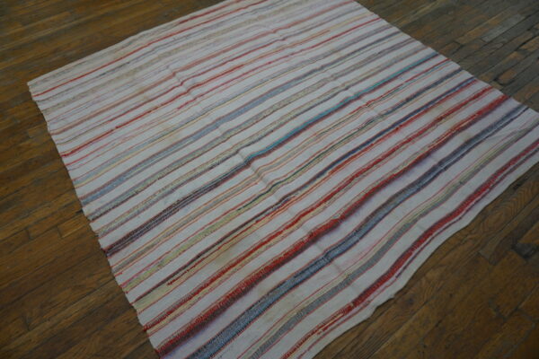 A flat-textured rug with cream, red, and blue stripes lies diagonally on an aged wood floor.
