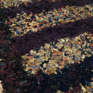 Highly textured rag rug featuring wide horizontal stripes of dark purple, black, maroon, and multicolored fabric scraps.