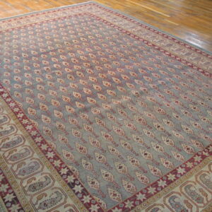 Ornate blue-gray rug featuring a repeating motif pattern and multiple red, cream, and paisley borders on hardwood.