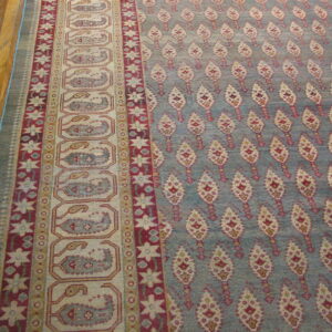 Low pile blue rug featuring ornate red and cream repeating patterns and layered borders on wood flooring.
