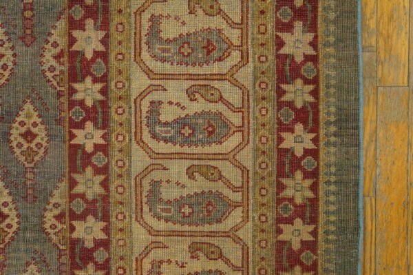 Traditional rug with red and tan geometric patterns, bordered by gray-blue stripes, resting on distressed wood flooring.