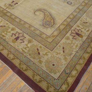 Beige and olive green rug featuring a paisley central medallion and wide floral border rests on distressed floorboards.