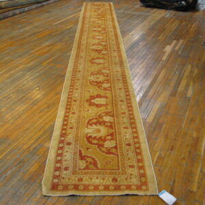 A beige and burnt orange runner rug lies on a shiny wood floor near rolls of stacked carpets.