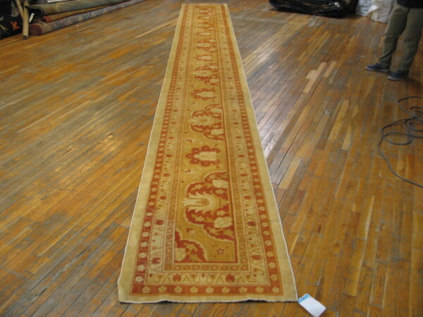 A beige and burnt orange runner rug lies on a shiny wood floor near rolls of stacked carpets.