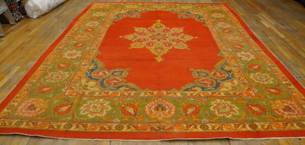 Large red medallion rug with wide olive green and multicolored borders resting on wood plank flooring.