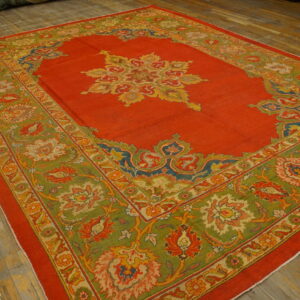 Vibrant red rug featuring a central medallion and wide green border with detailed floral patterns on wood flooring.