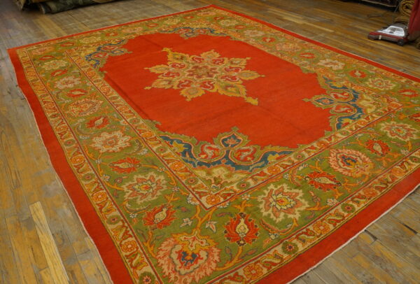 Vibrant red rug featuring a central medallion and wide green border with detailed floral patterns on wood flooring.
