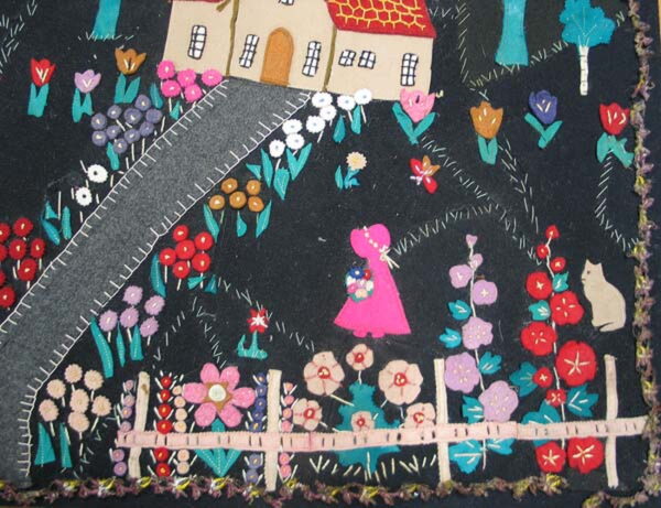 Colorful appliqué rug detail showing a stylized house, flower garden, pink figure, and cat on a dark ground.