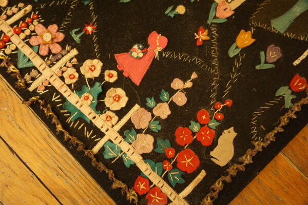 Black rug corner with colorful felt applique flowers, a pink figure, and hand-stitching on wood planks.