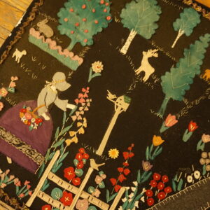 Black pictorial rug displaying bright appliqued figures, flowers, and trees on a wooden floor.