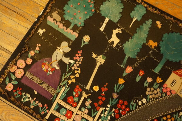 Black pictorial rug displaying bright appliqued figures, flowers, and trees on a wooden floor.