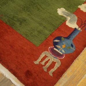 Rust red and green rug corner with a decorative vase and floral design lies on worn wood flooring.