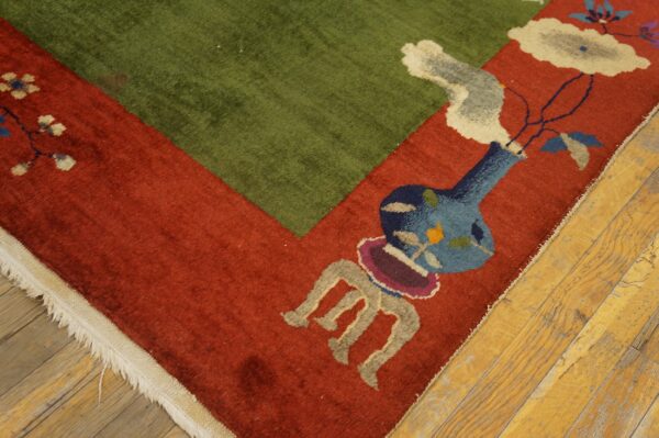 Rust red and green rug corner with a decorative vase and floral design lies on worn wood flooring.