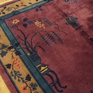 Plush maroon rug with a camel floral border and teal accents rests on light wood flooring.