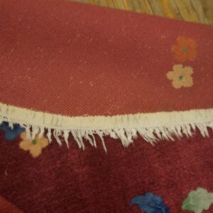 Detail of a rolled maroon rug showing fringe, woven backing, and multicolor floral designs on wood.