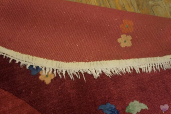 Detail of a rolled maroon rug showing fringe, woven backing, and multicolor floral designs on wood.