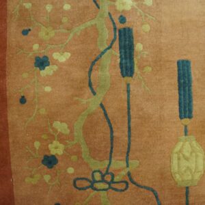 Detail of a salmon rug with stylized green branches, blue blossoms, and tassels on weathered yellow flooring.