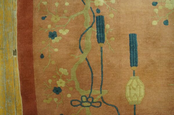 Detail of a salmon rug with stylized green branches, blue blossoms, and tassels on weathered yellow flooring.