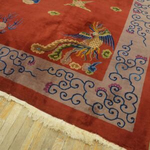 Rust-colored rug featuring a detailed colorful bird and scroll border lying on distressed wood planks.