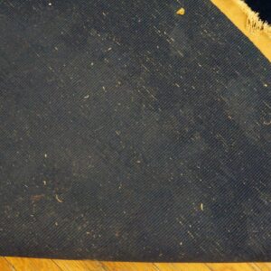 Dark blue woven rug backing with diagonal lines and a tan edge folds over light wood flooring.