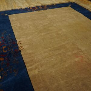 A large tan area rug features a thick blue border with intricate floral and gold scrollwork designs on a wood floor.