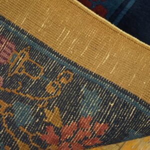 A curled navy rug exposes a woven tan backing and a patterned edge in gold, maroon, and blue.