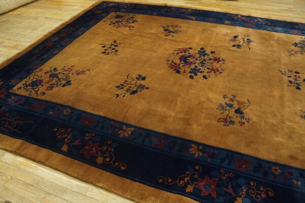 Medium-pile gold rug with dark blue floral borders and open pattern rests on worn hardwood flooring.