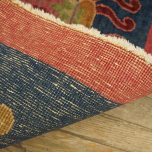 A rug is rolled back revealing its tightly woven red and blue foundation threads on rustic wood.