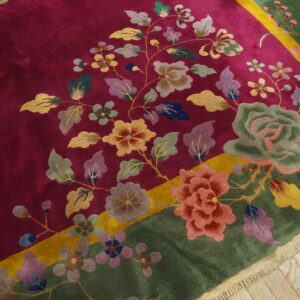 Plush magenta rug with multicolored floral design and wide moss green border rests on wood flooring.
