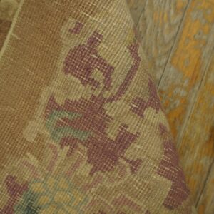 Rolled tan rug with low-pile texture and geometric burgundy pattern on chipped wood planks.