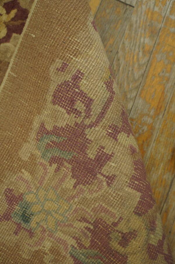 Rolled tan rug with low-pile texture and geometric burgundy pattern on chipped wood planks.