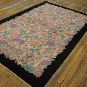 A low pile rug with a pink, purple, and blue floral pattern on a beige field framed by a thick black border, resting on worn wood floors.