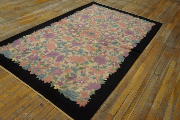 A low pile rug with a pink, purple, and blue floral pattern on a beige field framed by a thick black border, resting on worn wood floors.
